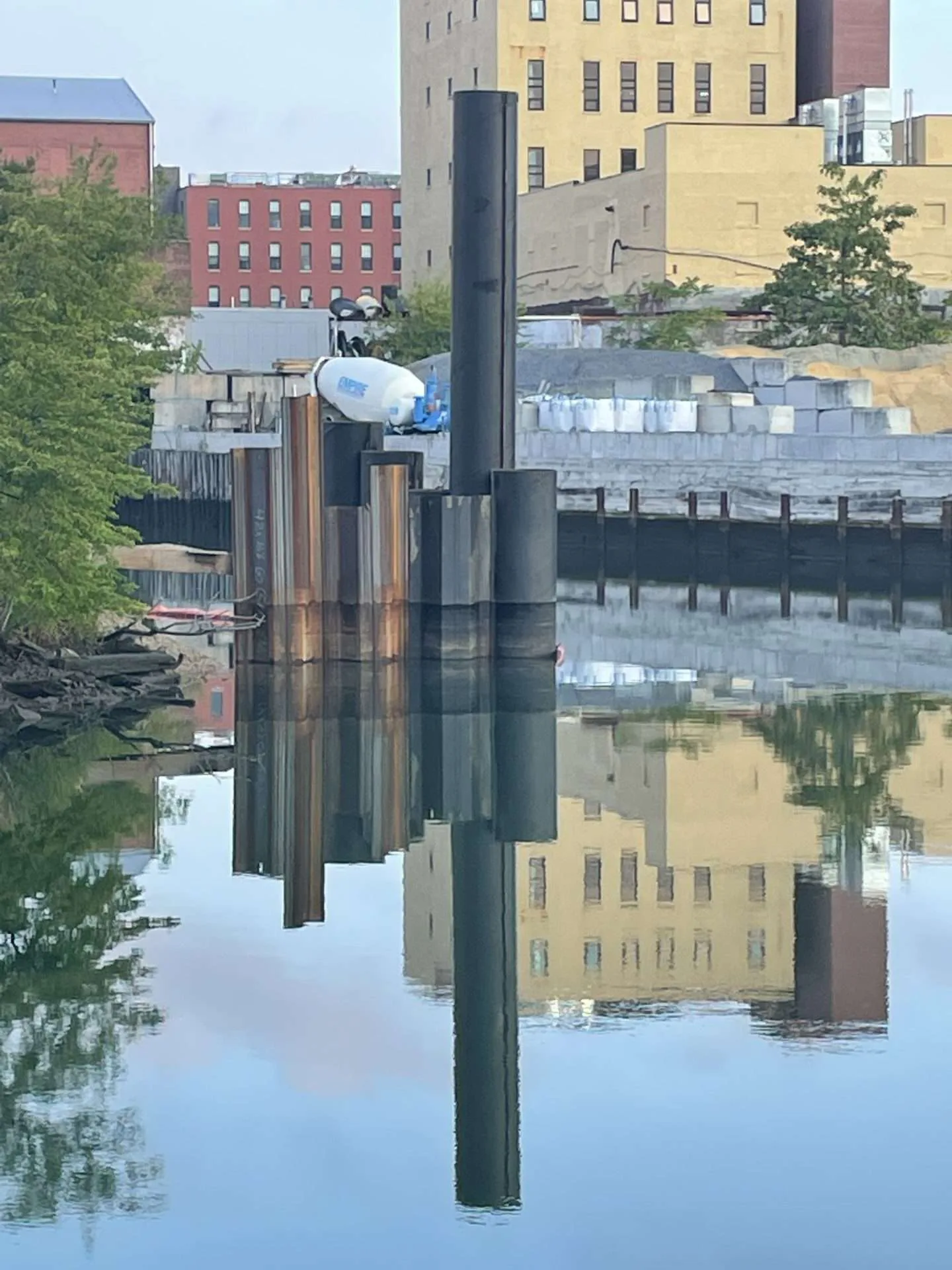 Photograph of urban waterfront with pristine mirror reflection: brick buildings, wooden pilings, and green trees reflected in still water below.