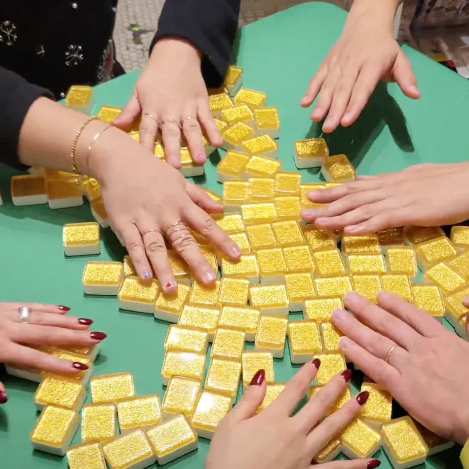Photograph of multiple hands with painted nails arranging small gold-colored cubic blocks on a green surface in a collaborative creative gesture.
