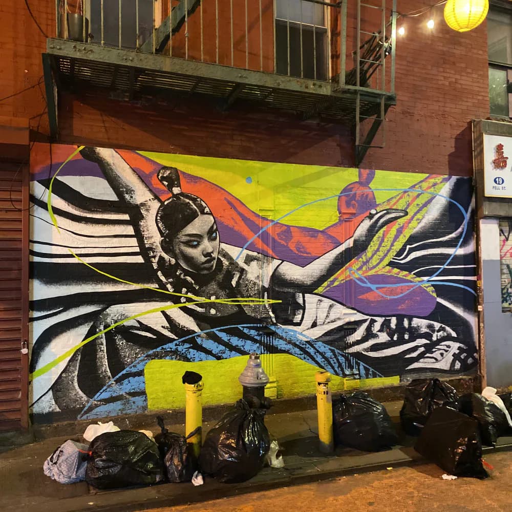 Vibrant mural by Gian Galang featuring a monochromatic figure with flowing hair against neon yellow, blue, red, and purple geometric shapes and line work on brick wall.