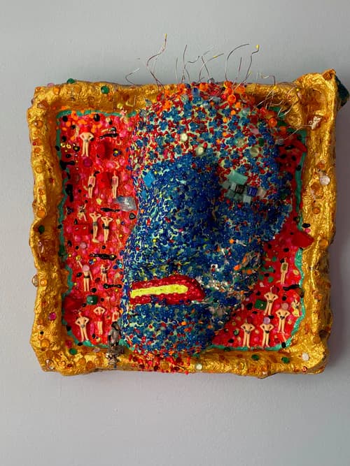 Mixed media sculptural face with sequins, tiny figurines, and colorful beading on gold framed background