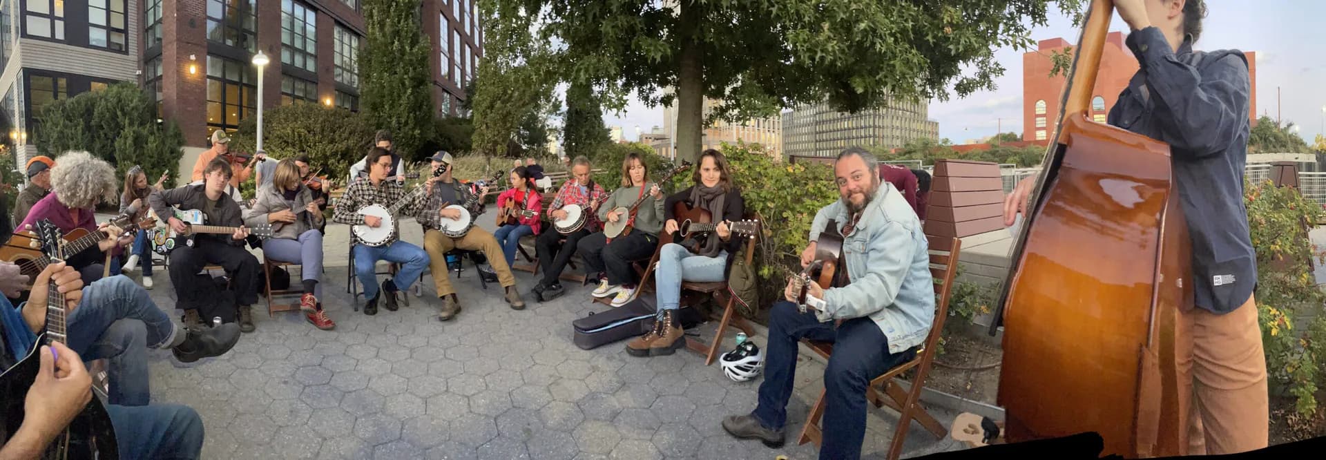 Panoramic photo of a large outdoor folk music jam session with banjos, guitars, and other instruments in an urban plaza