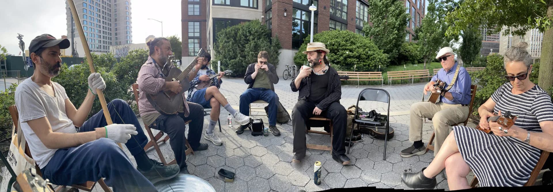 Panoramic photo of a small outdoor music gathering with musicians playing guitars, harmonica, and other instruments