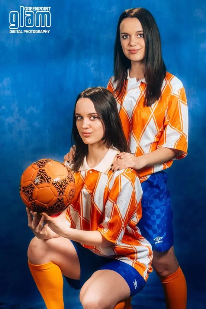 Portrait photograph by Greenpoint Glam: two women in matching orange and white diamond-patterned athletic jerseys, one kneeling holding decorative soccer ball, against blue background.