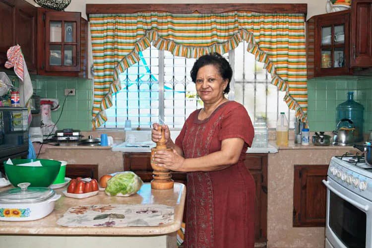 Woman in burgundy dress standing in a kitchen with green walls, holding a wooden pestle, striped yellow and blue curtains visible behind her.