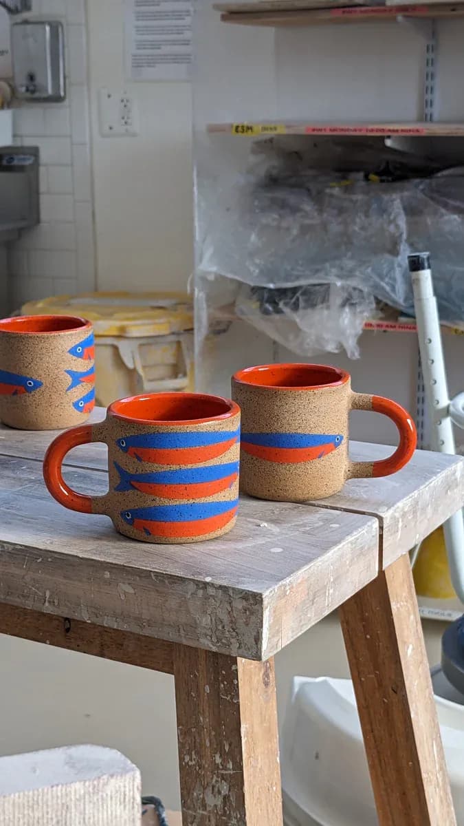 Three ceramic mugs with cork bodies, orange and blue striped hand-painted bands, and bright orange glazed interiors.