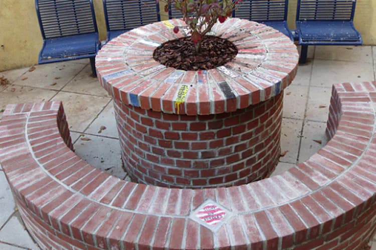 Circular brick planter with central vegetation, surrounded by curved brick seating, with blue chairs visible in background.