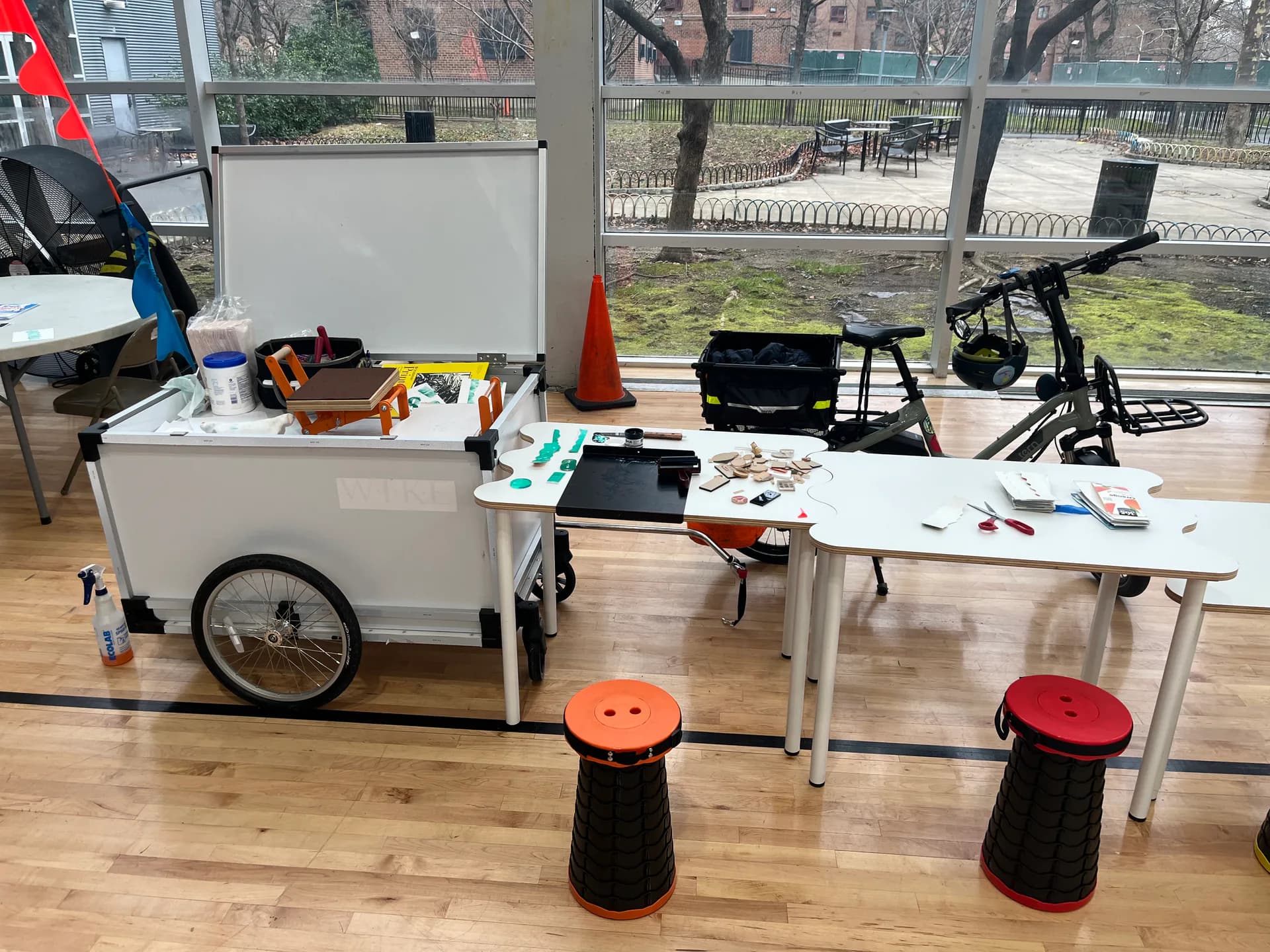 Interior studio space with large windows overlooking cityscape, white cart with supplies, work tables, and red/orange stools.