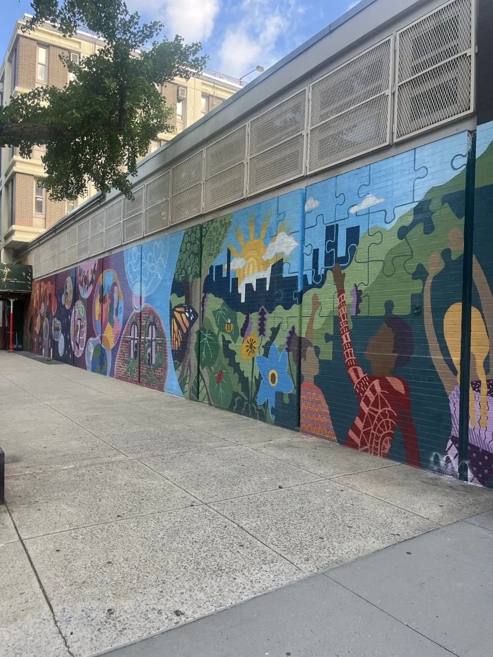 Large-scale mural featuring colorful puzzle-piece motifs, cityscape silhouette, sun, and nature elements in blues, greens, purples, and earth tones on an urban wall.