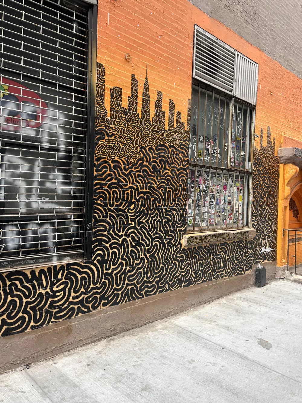 Mural with black and golden-yellow striped wavy pattern forming skyline shapes on a brick building facade; abstract line-work design.