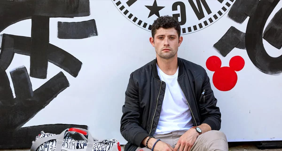 Man in black jacket and white shirt posing in front of a black and white graphic mural with geometric shapes and a red Mickey Mouse icon.