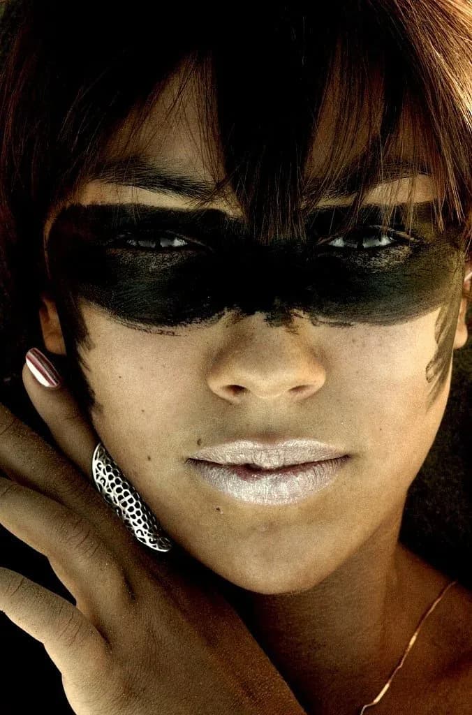 Close-up portrait of a person wearing dark oversized sunglasses and white lipstick, with brunette hair and dangling earrings against a black background.