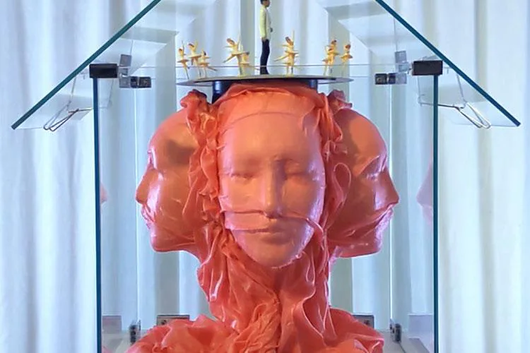 Red resin sculptural bust with multiple overlapping faces displayed in a clear acrylic case with overhead lighting.