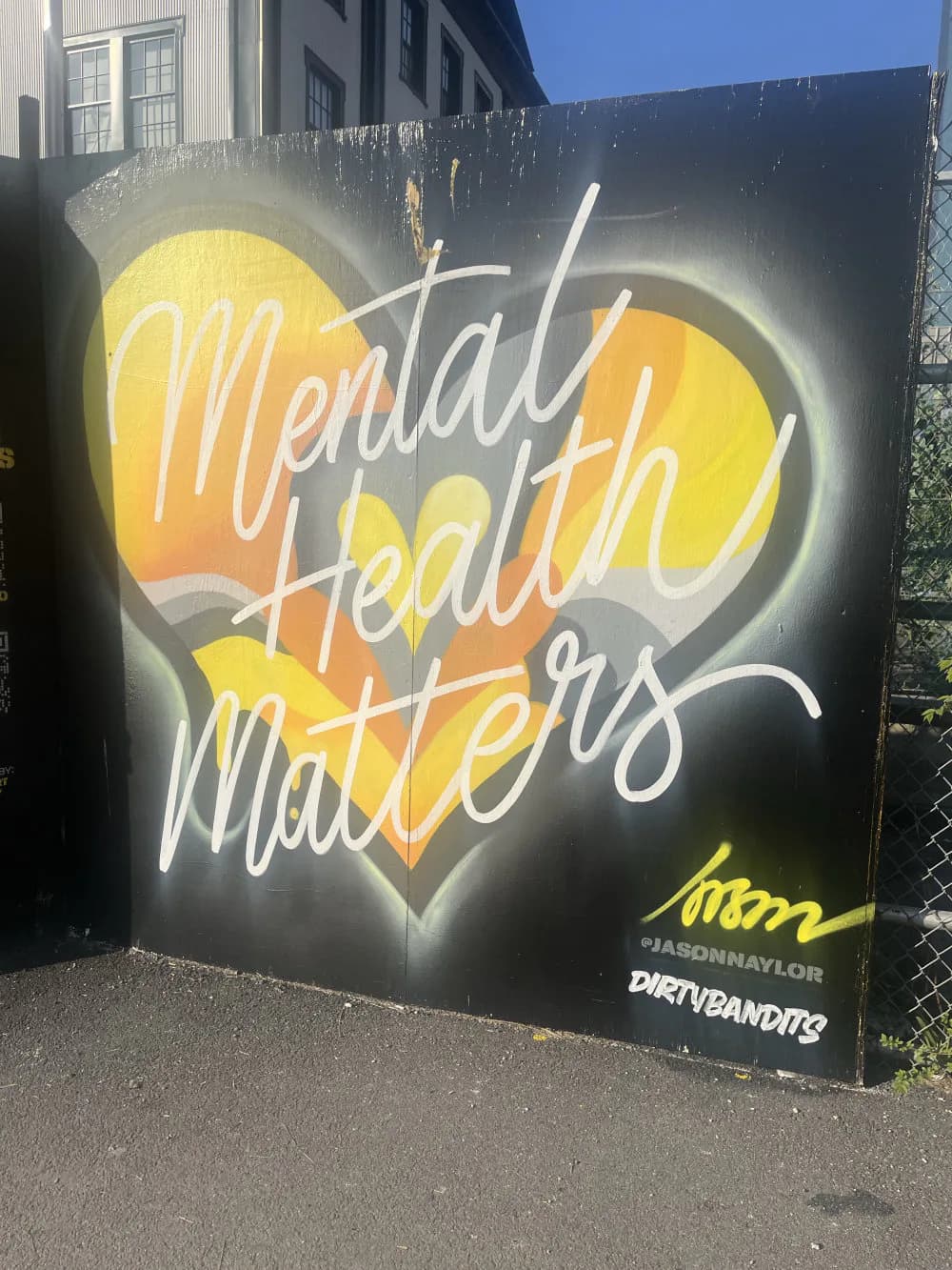 Neon sign mural featuring a yellow heart with white cursive text reading 'Mental Health Matters' against a dark background, signed by Jason Naylor.