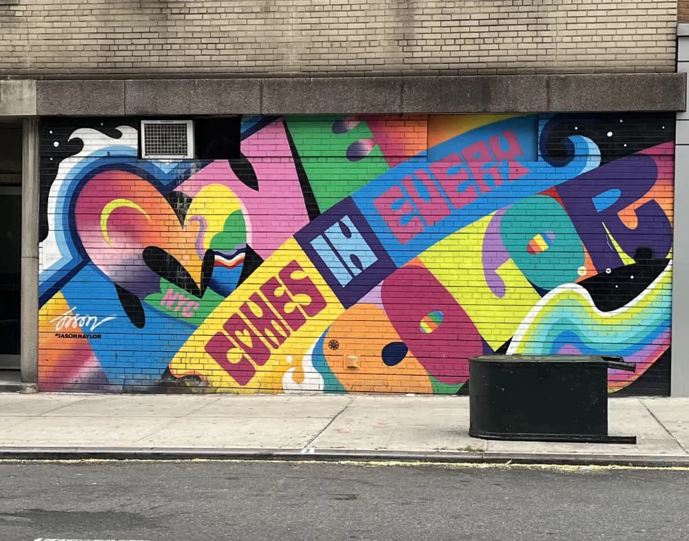 Vibrant spray-painted mural on brick with overlapping graffiti-style lettering in pink, blue, yellow, and green with abstract geometric shapes.