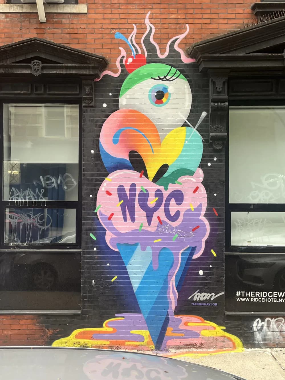 Vibrant mural depicting a whimsical ice cream cone with googly eye, colorful striped body, pink brain, and 'NYC' lettering, signed Jason Naylor.