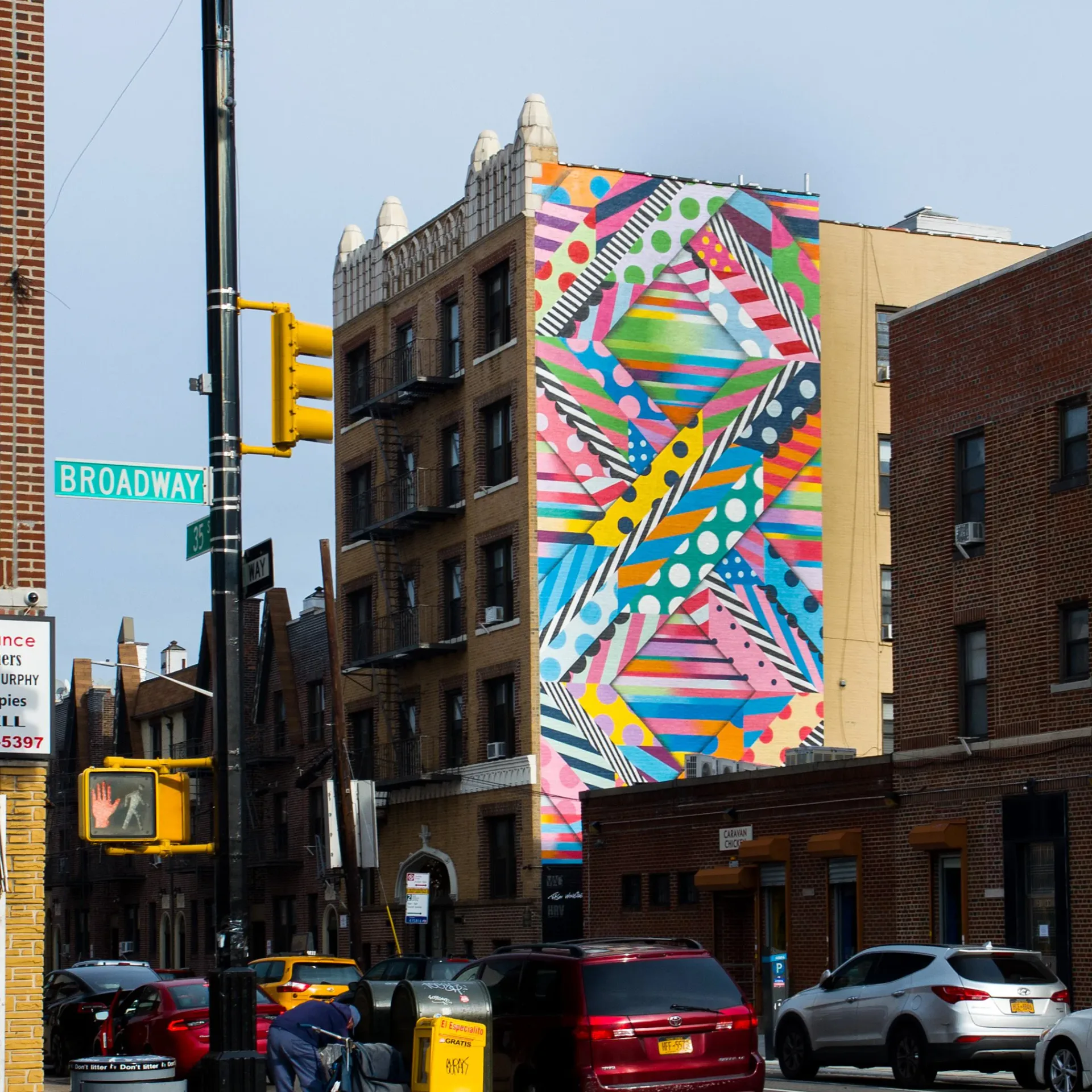 Urban street scene with large colorful geometric mural on building wall featuring rainbow stripes, patterns, and abstract shapes in bright colors.