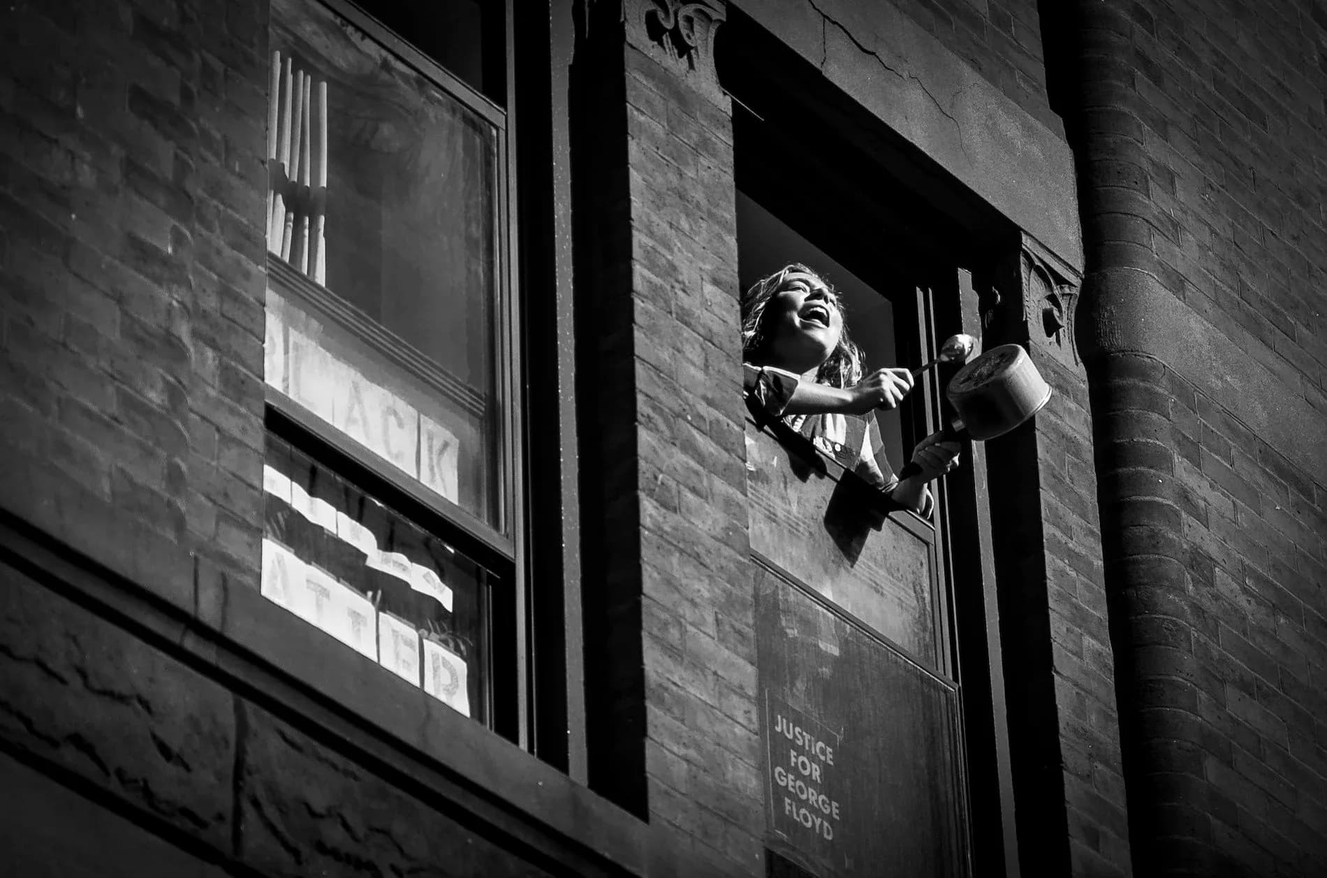 Laughing woman leaning out of a brownstone window banging a pot, Justice for George Floyd sign below