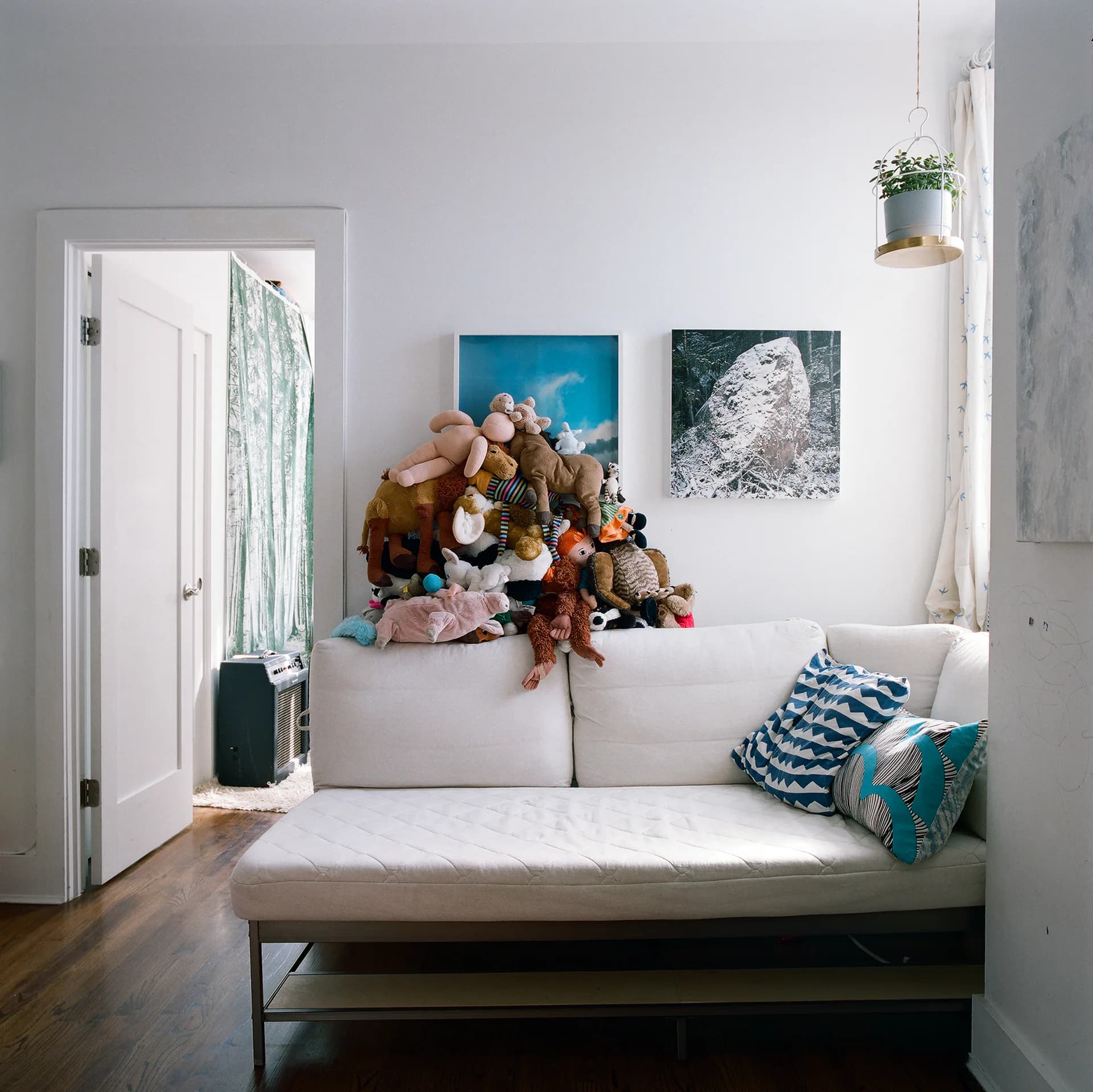 Living room interior with a large pile of stuffed animals on a white sofa, artworks visible on the wall