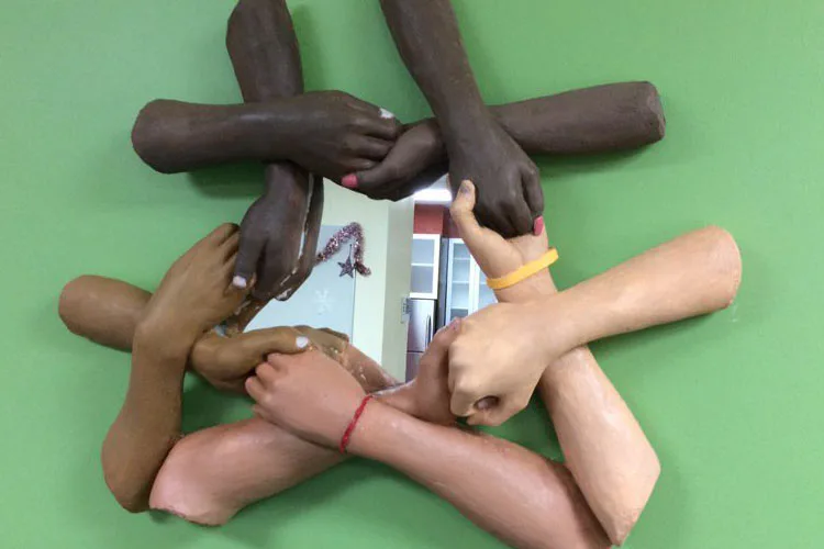 Colorful cast-resin sculpture of multiple hands of different skin tones arranged in a circle around a mirror, symbolizing unity and diversity.