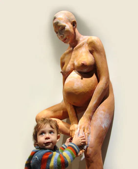 Polished bronze sculpture by John Ahearn depicting a pregnant woman cradling her belly, with a small child holding her leg, rendered in warm golden-brown tones.