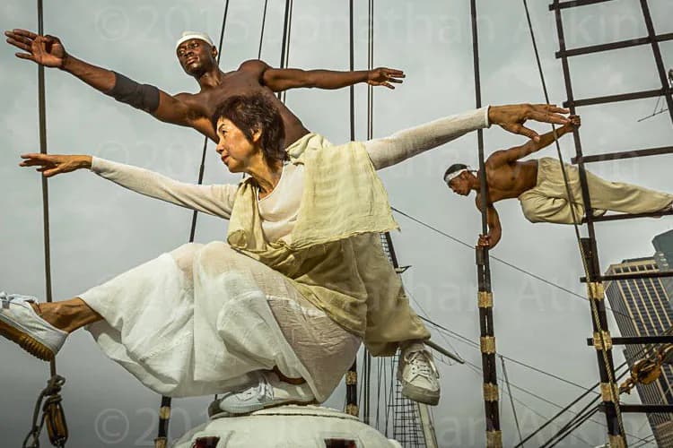 Suspended figures in yellow and white clothing pose dynamically against industrial rigging and sky, creating a theatrical aerial composition.