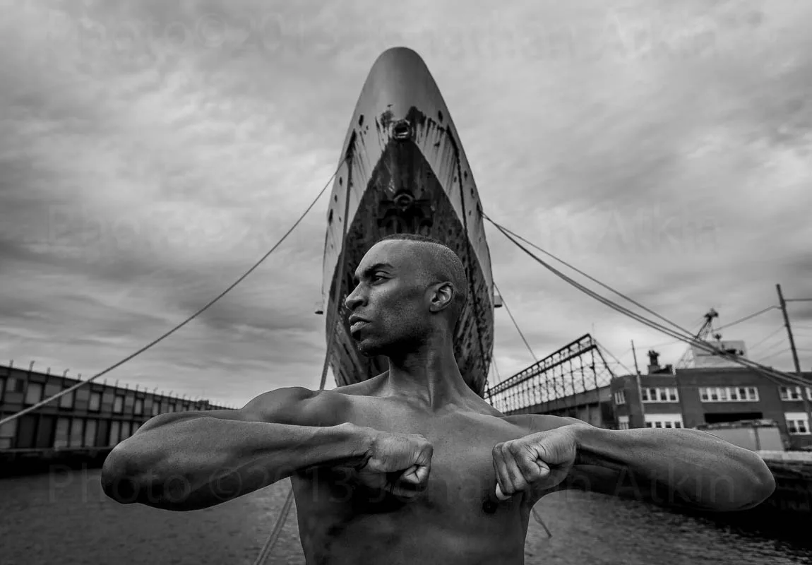 Black and white photograph of a monumental bronze sculpture depicting a figure with outstretched arms against stormy sky and industrial waterfront.