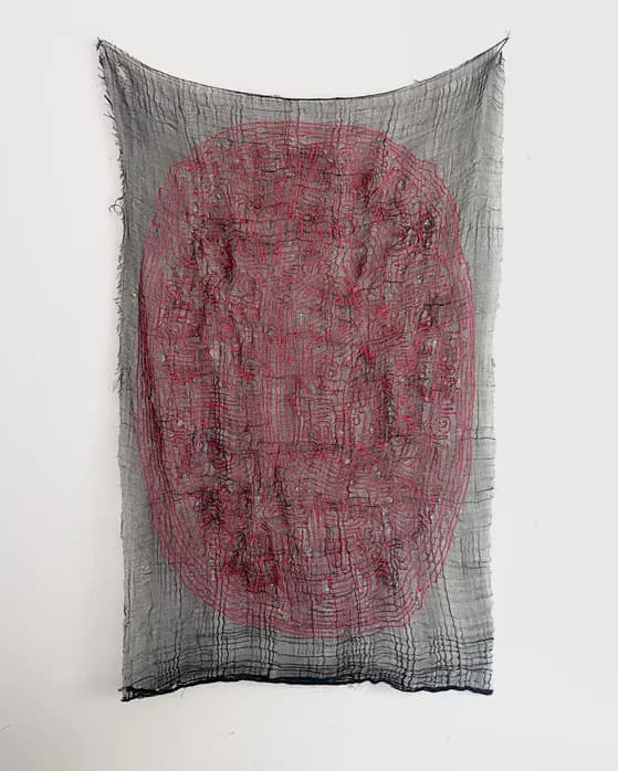 Gray mesh textile draped with burgundy-red dense woven section in center, creating three-dimensional form with textured overlay and fringed bottom edge.