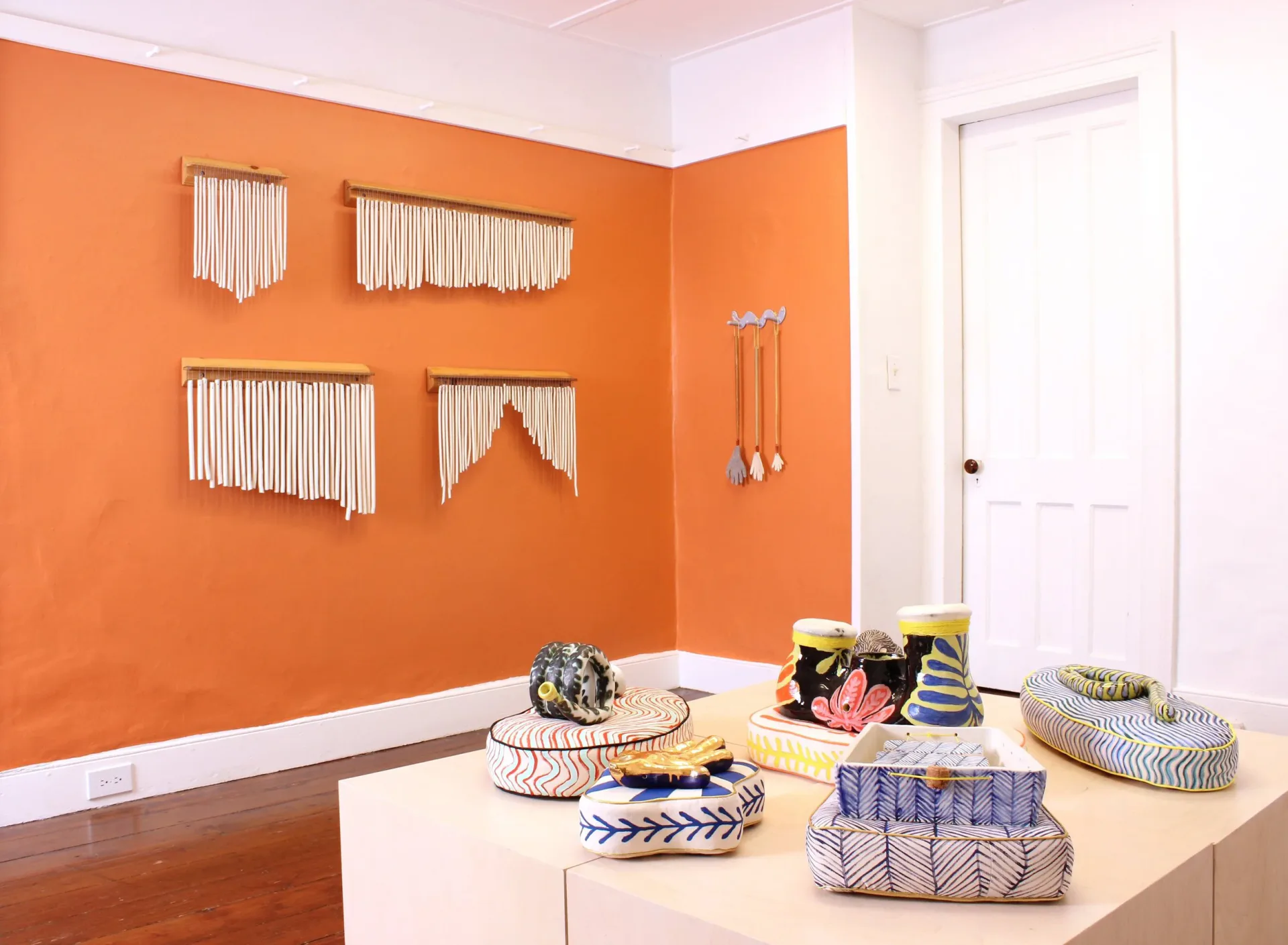 Gallery installation view featuring four ceramic wall-hung fringe pieces on an orange wall alongside whimsical hand-shaped hanging sculptures, with a plinth displaying colorful patterned ceramic vessels and boxes in the foreground