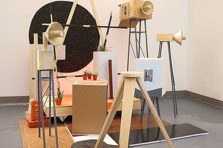 Mixed-media installation with cardboard forms, wooden structures, metal elements, and a large black circular backdrop with geometric brass accents.