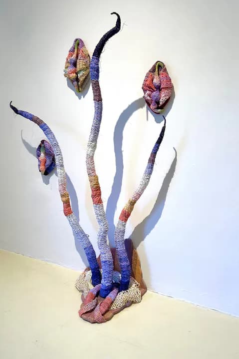 Three sculptural forms resembling stylized flowers or seed pods wrapped in multicolored thread in purple, blue, pink, and gold gradients.