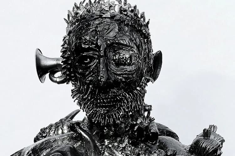 Black sculptural head with textured, spiky surface resembling metal or cast material; features trumpet protruding from mouth, exaggerated facial features, and decorative shoulder armor against white background.