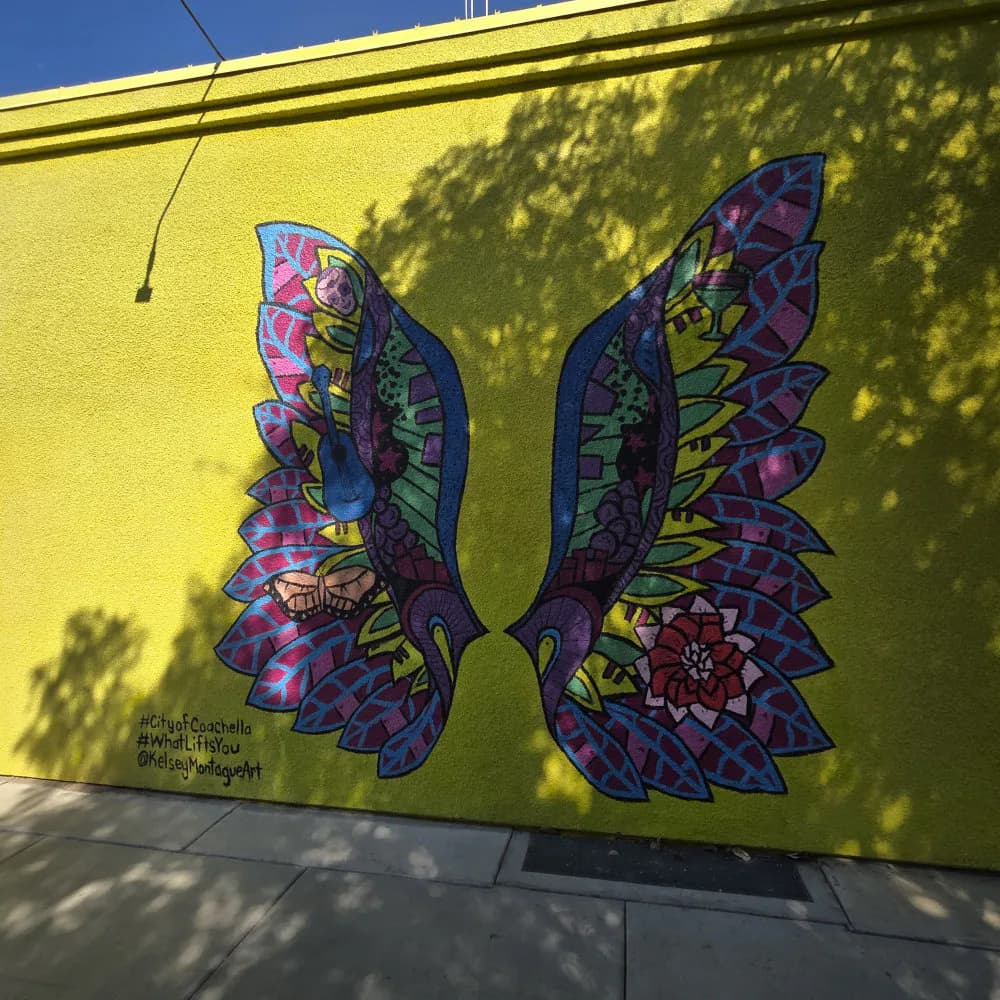 Mural on yellow wall featuring two symmetrical ornate butterfly wings in purple, pink, blue, and green with intricate geometric patterns and mandala-like designs.