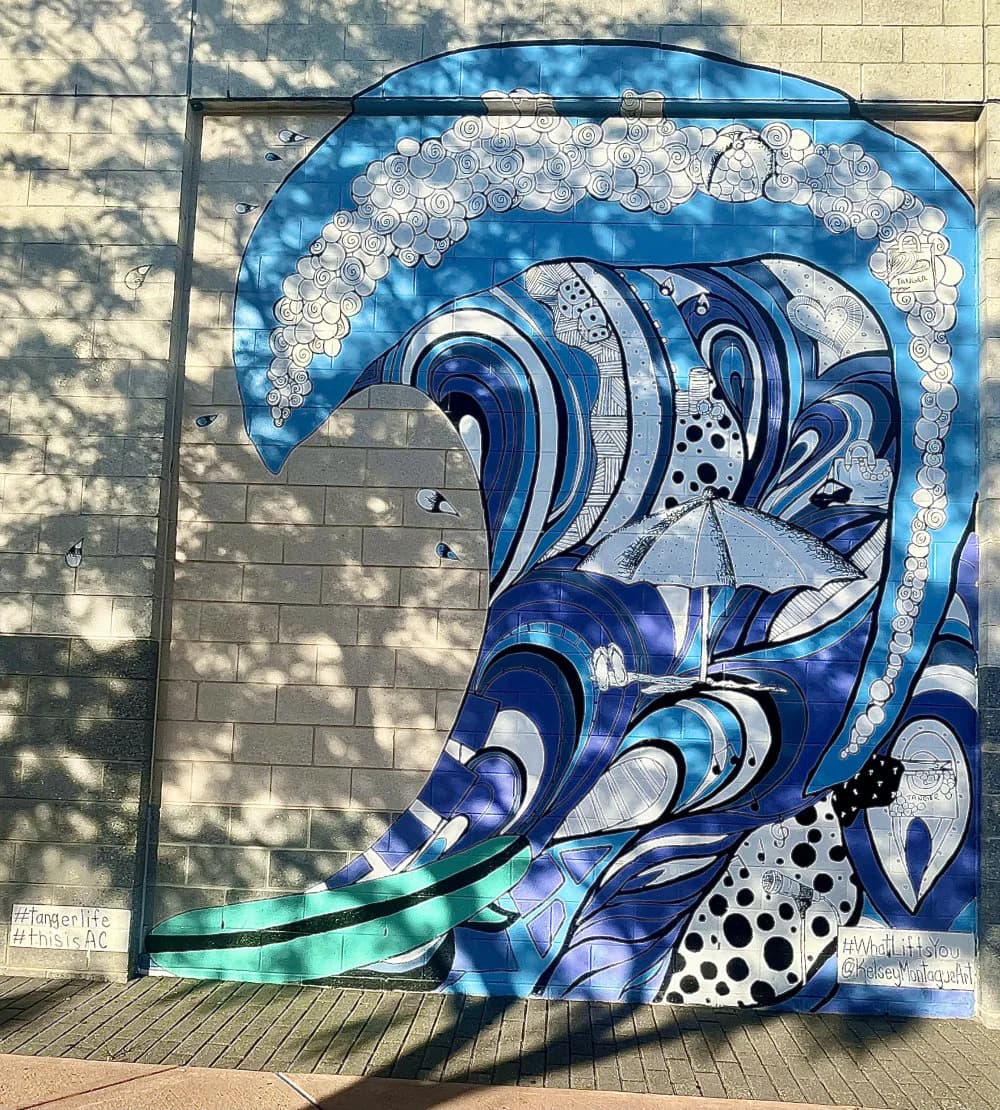 Large-scale mural of stylized blue wave with white clouds and geometric patterns, featuring flowing water forms and dotted textures on tan building wall.