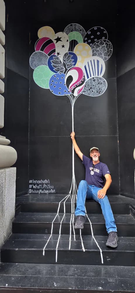 Mural on black wall showing a delicate stem holding a bouquet of patterned circles in various colors and textures, with artist posing below. Kelsey Montague's work.