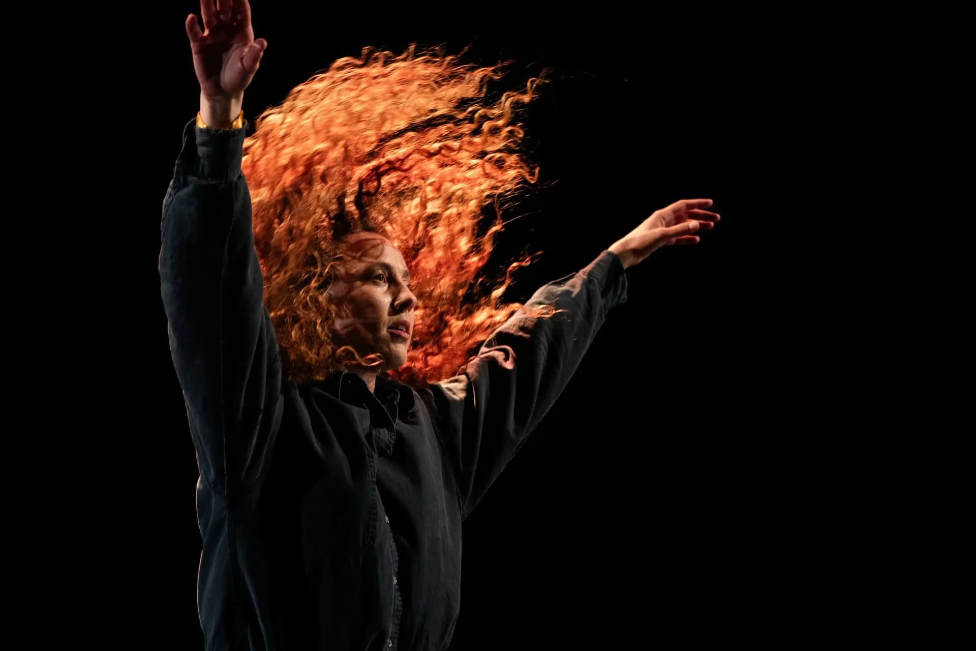 Performer with fiery red curly hair raised arms against black background