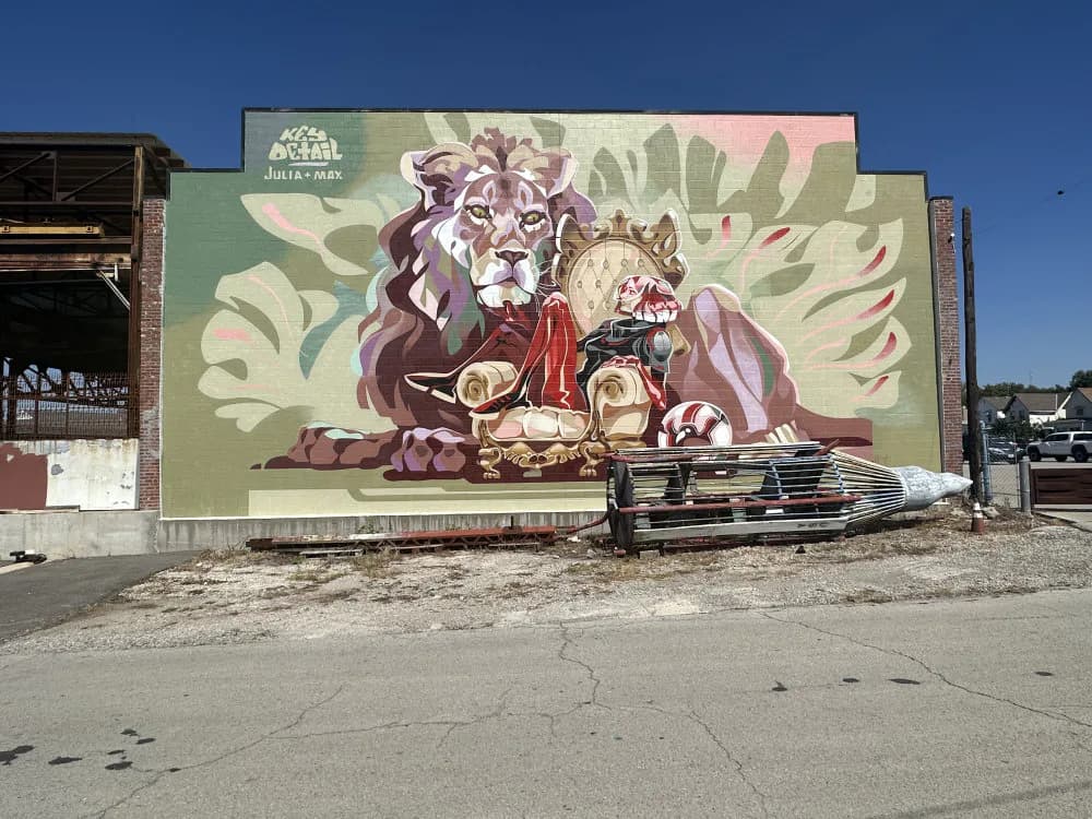 Large mural depicting a reclining figure with animals and decorative floral elements in muted greens, purples, and earth tones on an industrial building.