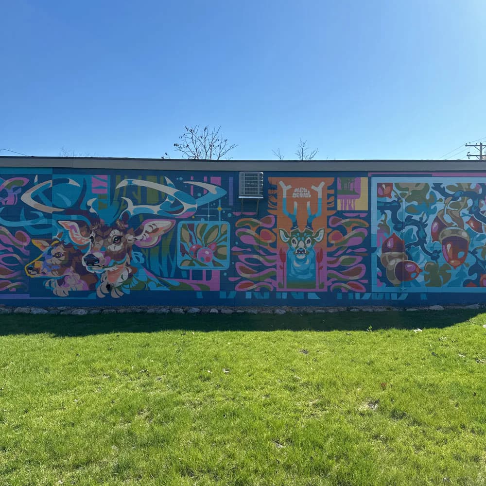 Horizontal mural with vibrant blue, purple, orange, and teal abstract creatures and figures spanning the length of a building against green grass.