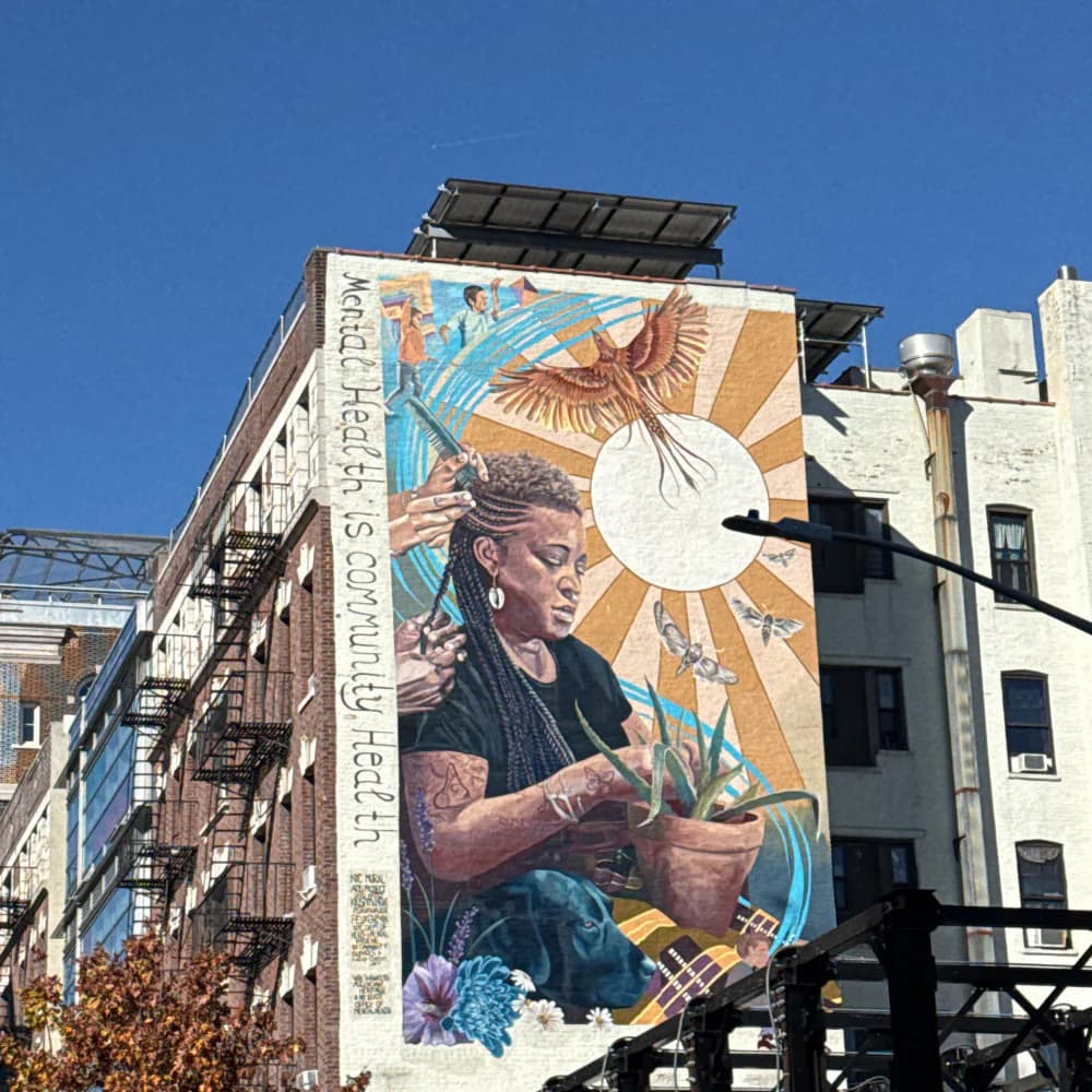 Large-scale mural on urban building exterior showing a person with tattoos tending to plants, with blue sky and golden sun rays.