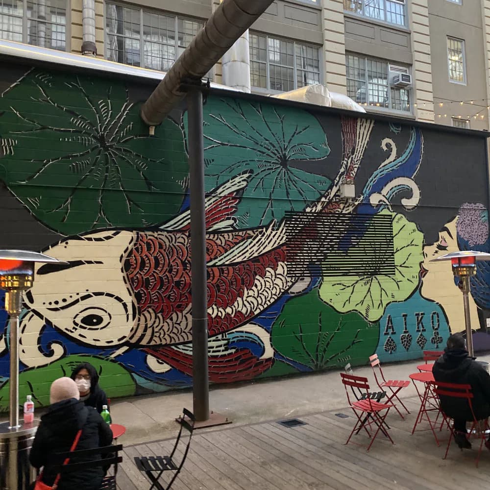 Outdoor mural by Lady Aiko featuring stylized fish, lotus flowers, and decorative patterns in green, cream, navy, and red on an urban wall.