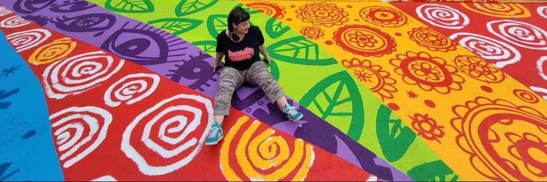 Vibrant painted floor installation in red, purple, green, and orange with concentric circular patterns; person sitting on colorful surface.