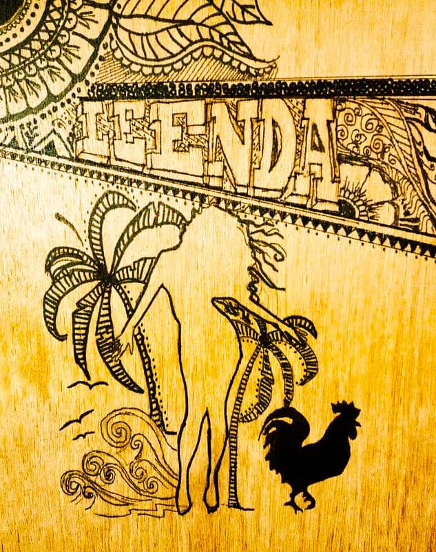 Woodburned illustration on light wood showing woman with plantains, rooster, and ornamental border with "FENDA" text.