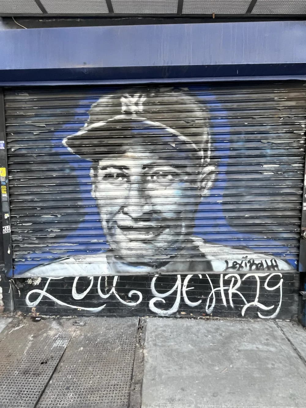 Monochromatic portrait mural on a shuttered storefront showing a man's face with a cap, painted in white and blue tones with graffiti text below.