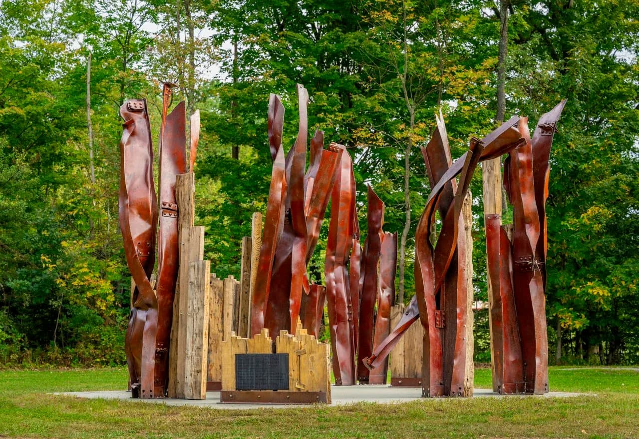 Outdoor sculptural installation featuring tall wooden forms in deep red and burnt sienna, arranged in a grove with a plinth base.