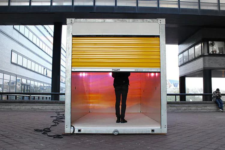 Mixed-media installation by Lionel Cruet featuring a glass and metal box with yellow horizontal slats above and a warm gradient red-orange panel below, silhouette of standing figure visible inside, positioned in urban plaza.