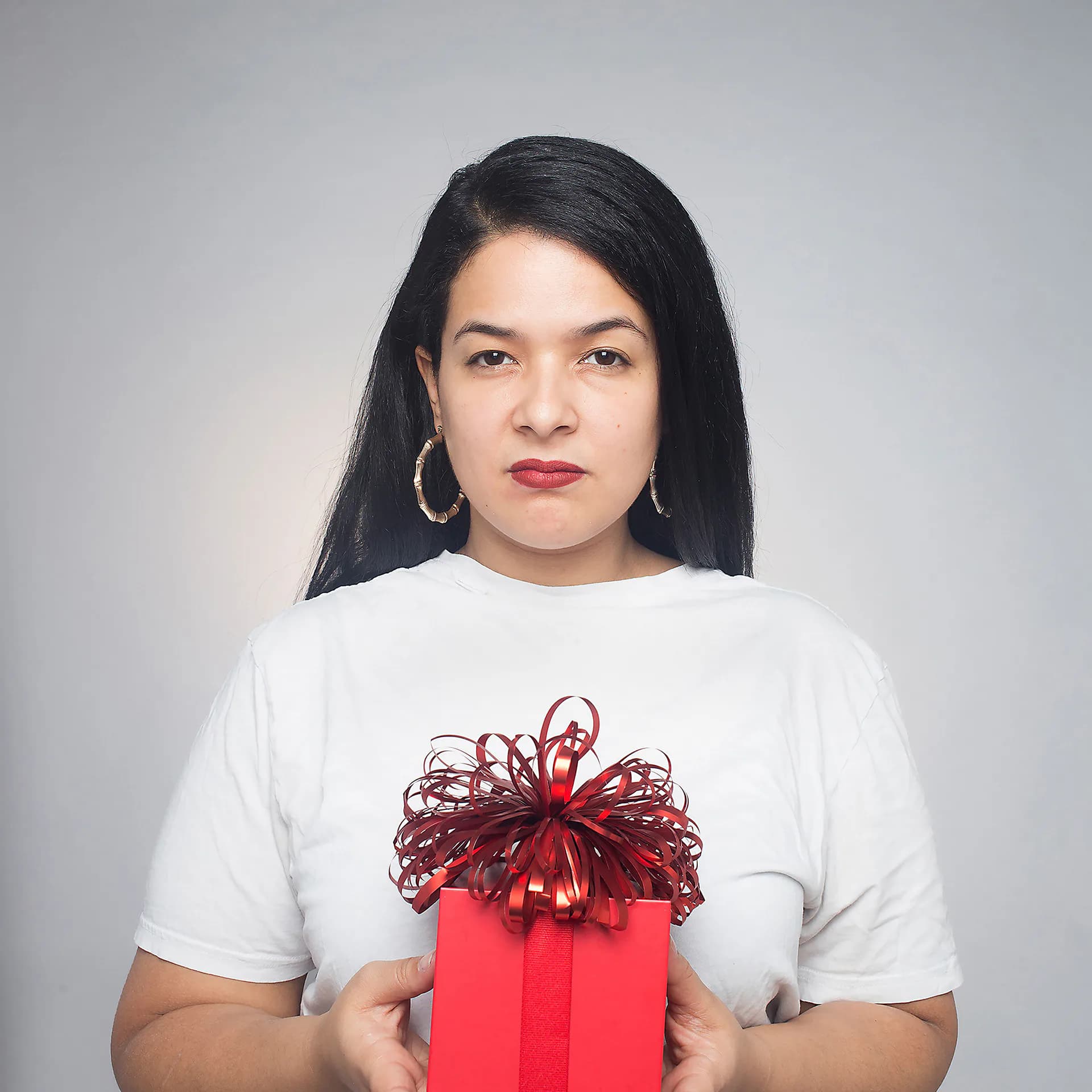 Portrait photograph of a woman in white t-shirt holding a red gift box with oversized burgundy ribbon bow against neutral gray background.