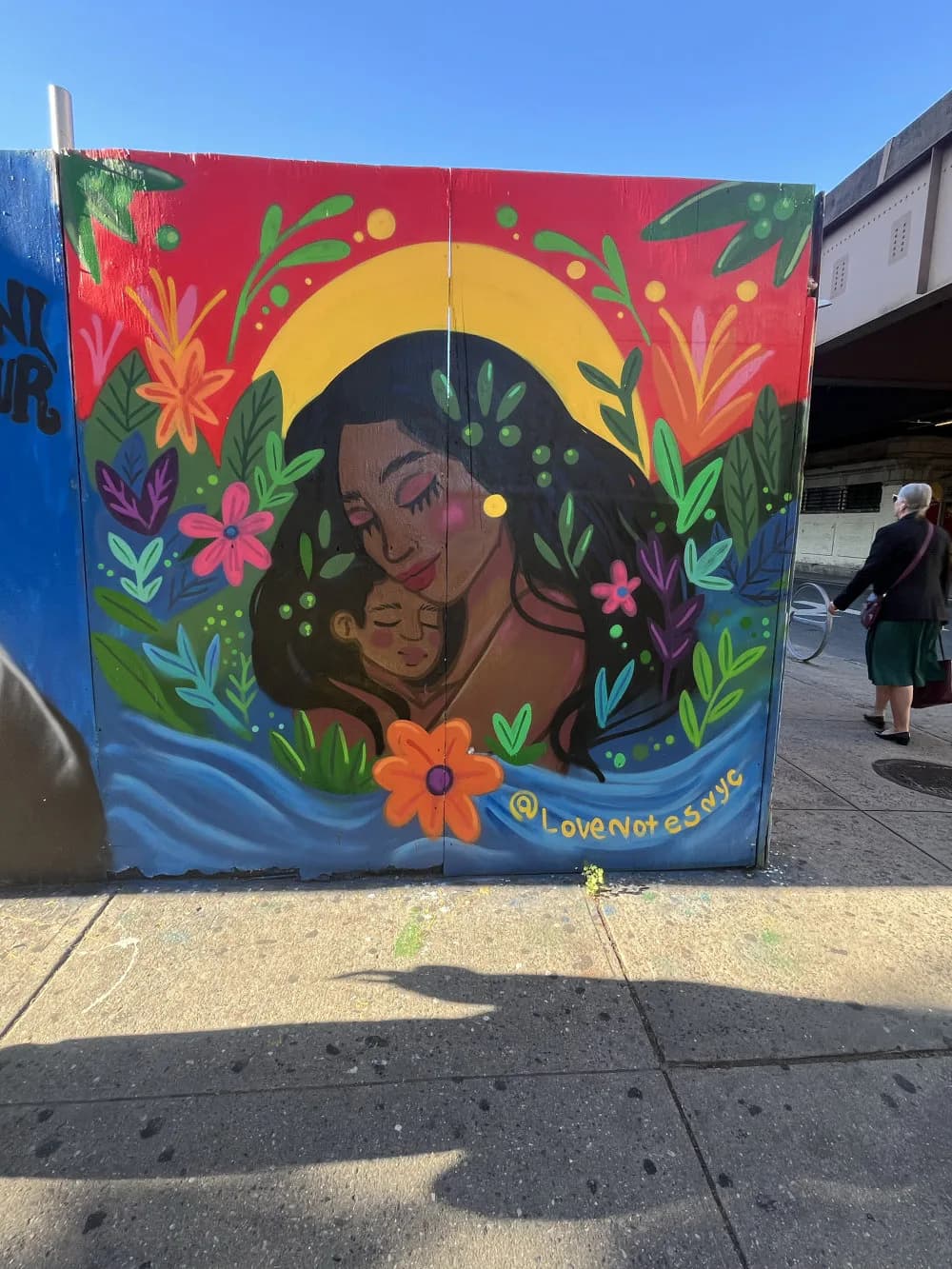 Colorful mural by LOVENOTES depicting a woman and child surrounded by vibrant flowers, plants, and a golden sun on a red background with blue water at base.