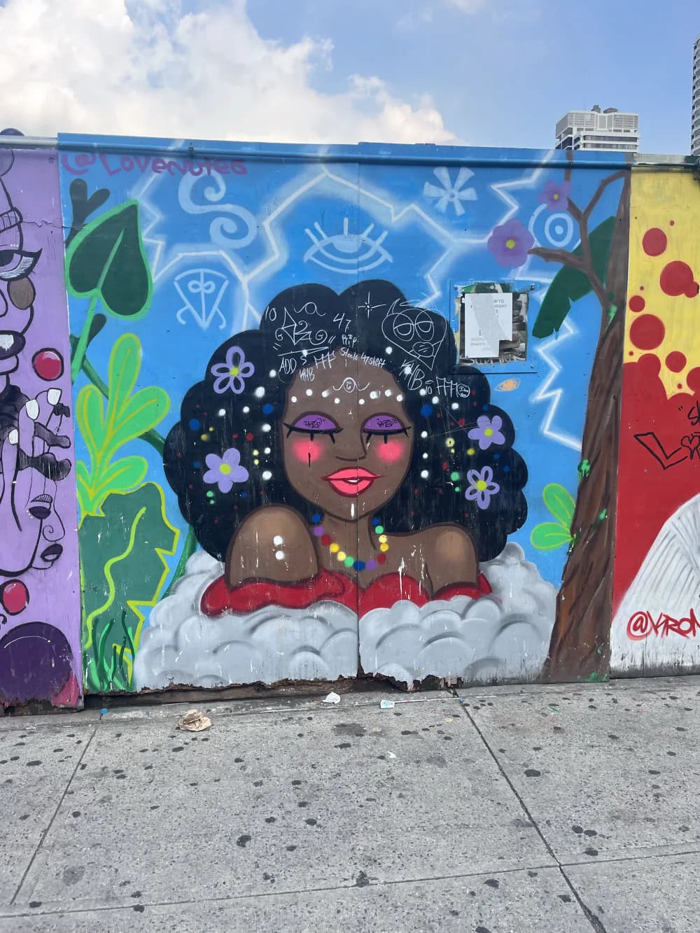 Street mural featuring a dark-skinned figure with black curly hair, purple lips, and white dots, surrounded by green foliage and celestial symbols on blue background.