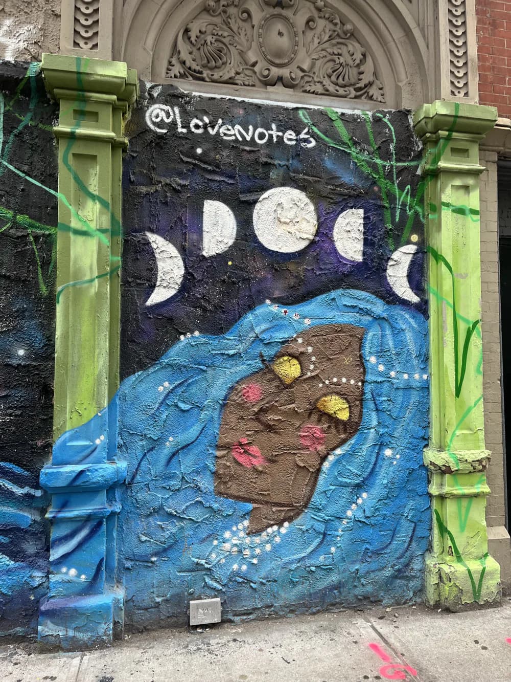 Mixed-media mural with moon phases at top, blue flowing fabric or water, brown textured form, and colorful dots, framed by lime-green architectural columns.