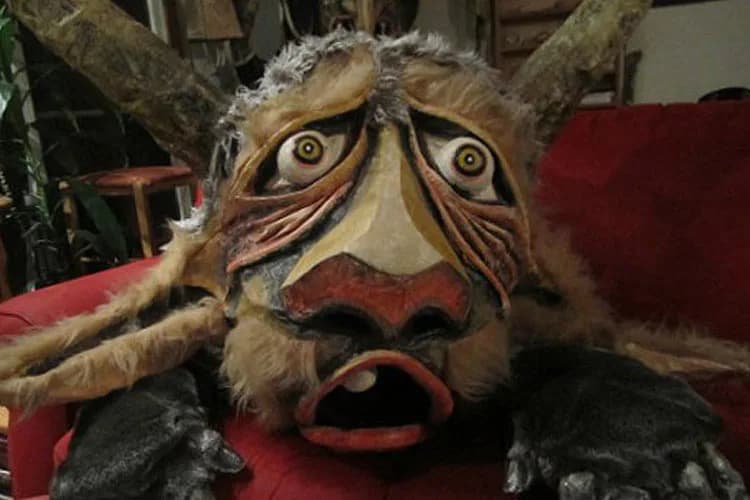 Wooden sculptural mask with expressive carved face, wide eyes, open mouth, and white/cream painted details with brown striations and dark fur or fiber accents.