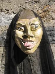 Golden-hued sculptural mask with black veil, stylized facial features including elongated eyes and protruding tongue, mounted on dark fabric.